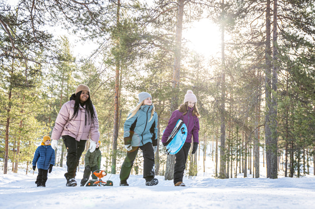 Viisi nuorta kävelee talvivaatteet päällään lumisessa metsässä ja kantavat erilaisia lumileikkivälineita, kuten stigaa ja pulkkaa.